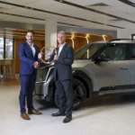 Dos hombres en trajes sostienen un trofeo junto a un coche moderno.